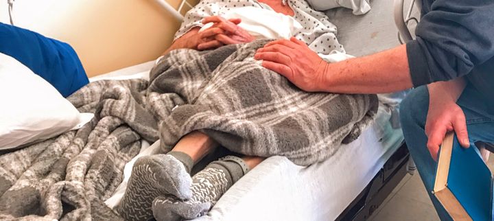 One man visiting another in a hospital bed. He holds a book and touches the other gently on the knee.