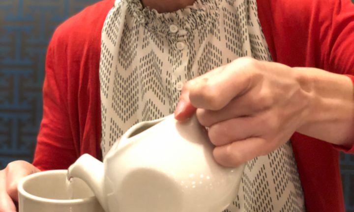 Photo of a woman in a vermillion sweater pouring hot water from a china teapot into a cup.
