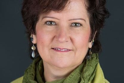 Portrait of a while woman with brown hair wearing a green jacket, green scarf, earrings and a necklace.